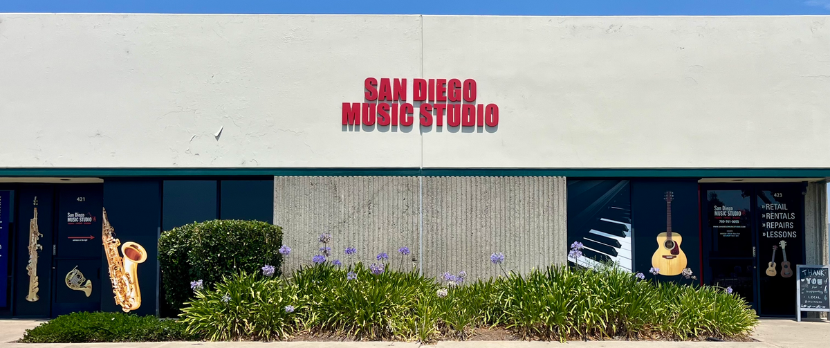 Front exterior of a music studio building with large red 'SAN DIEGO MUSIC STUDIO' lettering on a white facade, dark windowed storefronts, decorative saxophone and guitar posters, and landscaped shrubs in front.