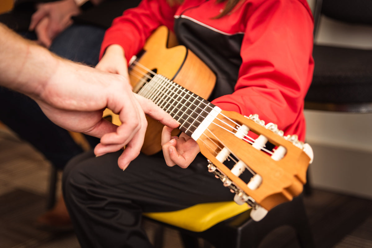 A close-up lifestyle shot of a person playing a classical guitar, showing their hands on the fretboard and headstock, wearing a red and black tracksuit while sitting on a yellow chair.