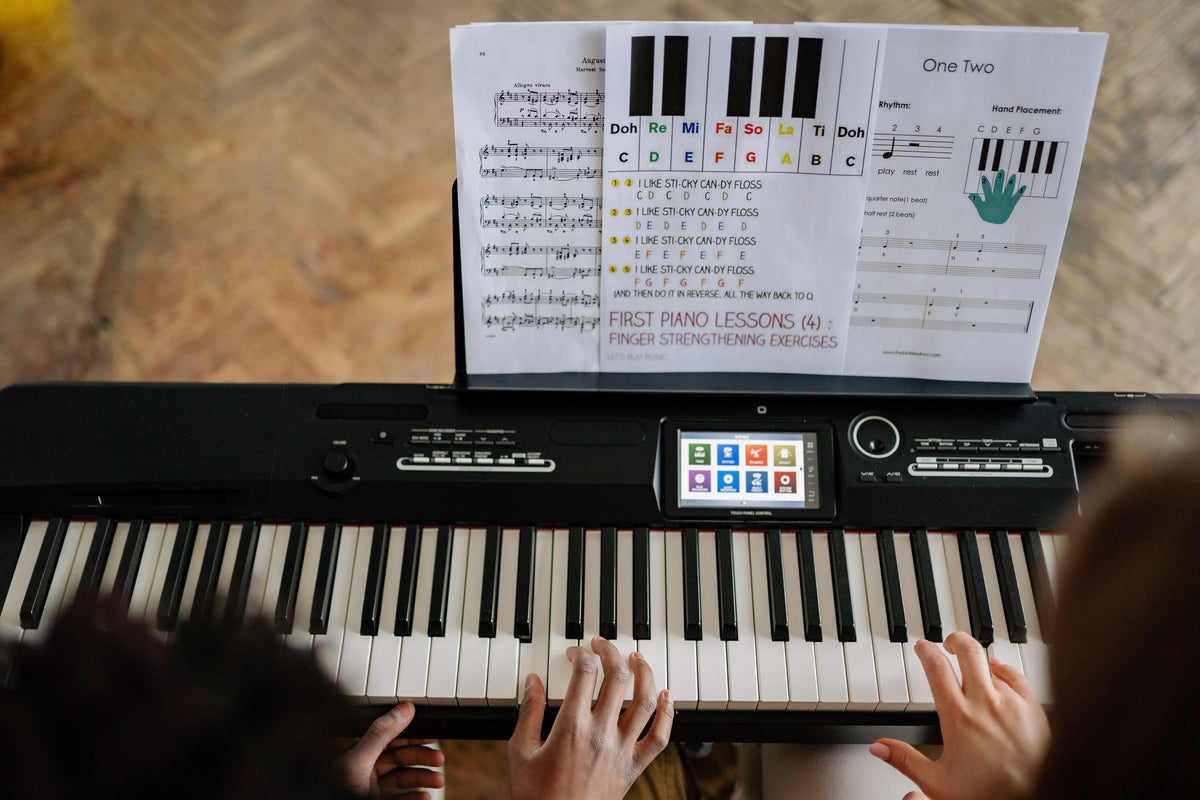 A person's hands playing a black digital piano keyboard, with a printed piano lesson sheet displayed above the keys.