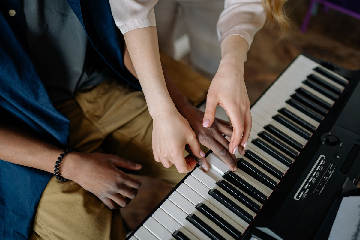 Close-up of two people's hands playing a black digital keyboard, with one person wearing a blue vest over a gray shirt and tan pants, and the other wearing a white long-sleeve top.
